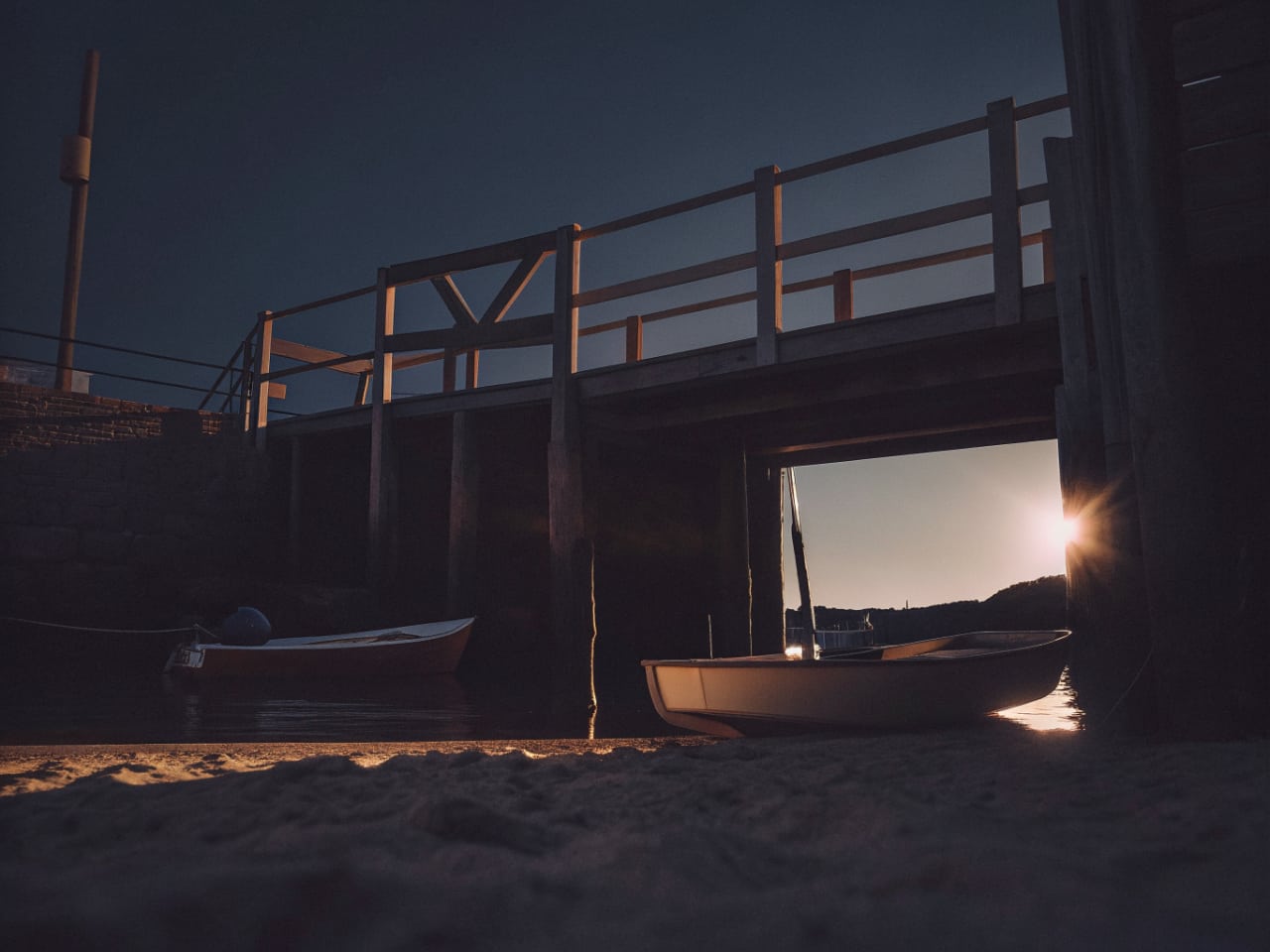 Photorealistic quiet small harbor at sunset seen from a sandy shore, an empty weathered wooden jetty leading out over ca